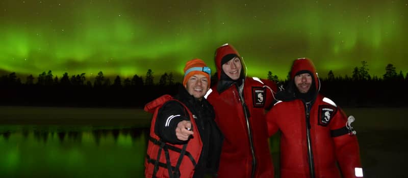 three people in red suits by a lake under green aurora (small logo on suits — check)