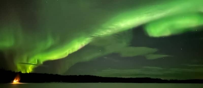 green aurora arcs over a lake with campfire glow on the shore