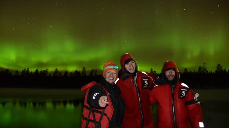 three people in red suits by a lake under green aurora (small logo on suits — check)