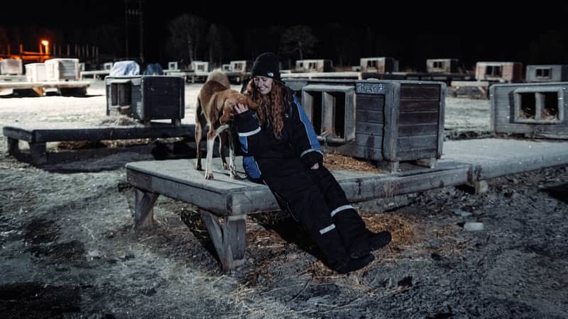 Mujer con traje térmico sentada junto a un husky sobre la plataforma de una caseta, bajo focos de noche