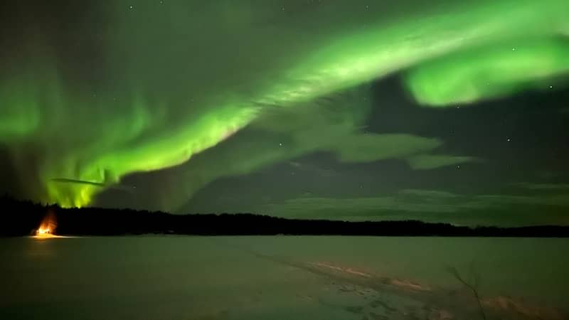 green aurora arcs over a lake with campfire glow on the shore