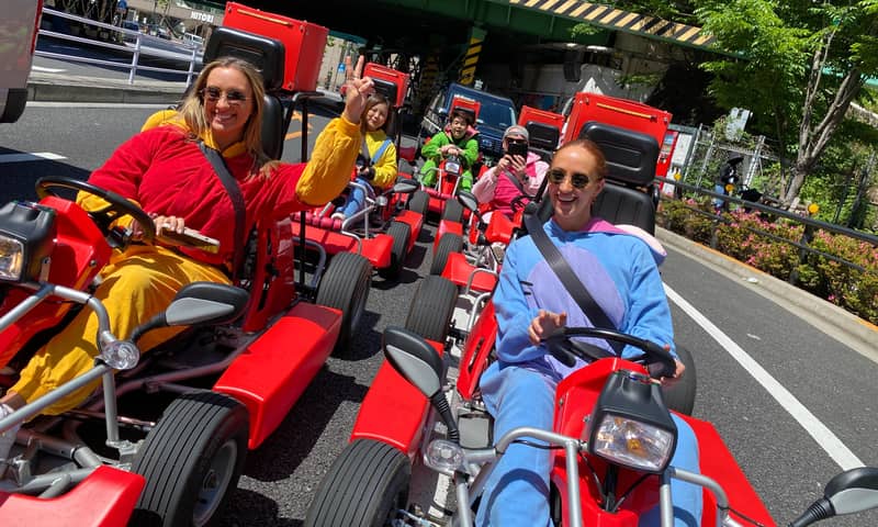 Day, wide street with trees, group of red karts, drivers in yellow/blue onesies waving to camera