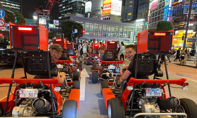 Night, red karts with rear cargo boxes lined up under neon signs, drivers seen from behind