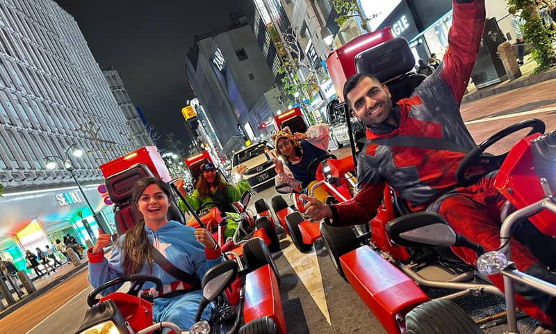 Night, woman in blue-green onesie and man in red at the wheel, neon Shibuya street, motion