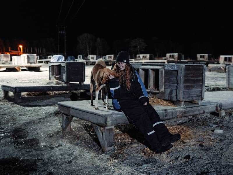 Woman in thermal suit sits with a husky on a dog-house platform under floodlights at night