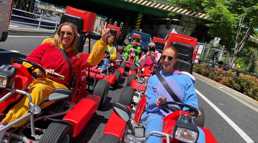 Day, wide street with trees, group of red karts, drivers in yellow/blue onesies waving to camera