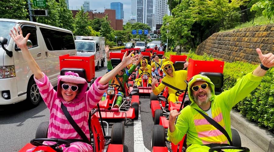 Day, group in pink/green striped onesies in red karts, arms up, on the road
