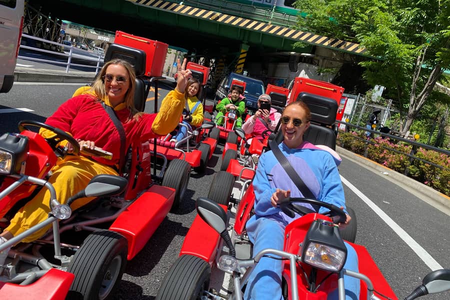Day, wide street with trees, group of red karts, drivers in yellow/blue onesies waving to camera