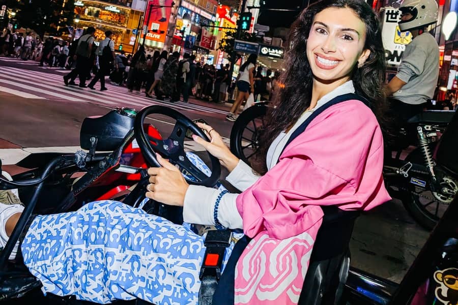 Day, woman in pink jacket at the wheel smiling, Shibuya crossing crowd behind