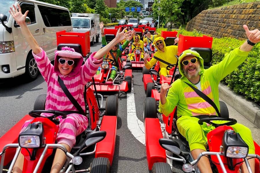 Day, group in pink/green striped onesies in red karts, arms up, on the road