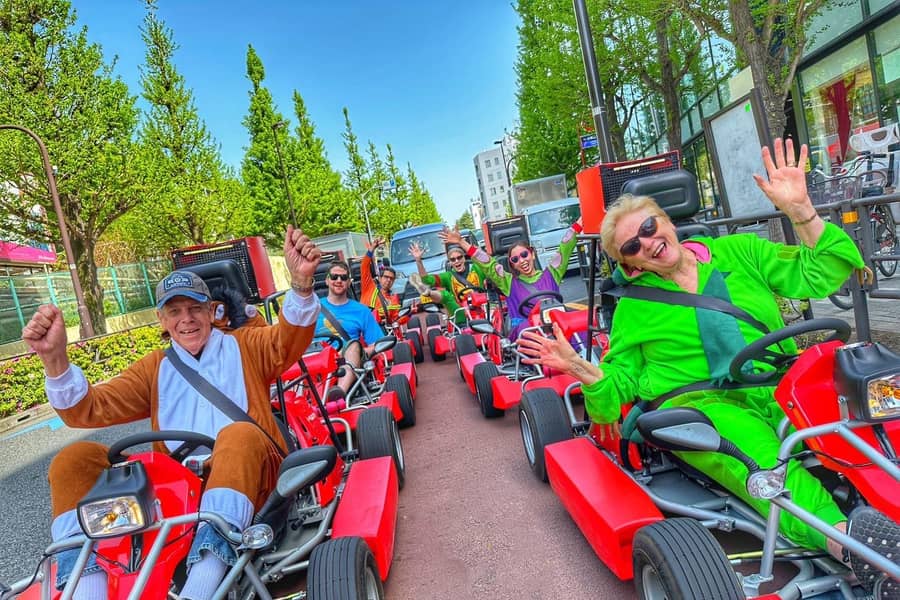 Day, wide avenue, ~8 red karts in a line, bright costumes, arms up, spring greenery