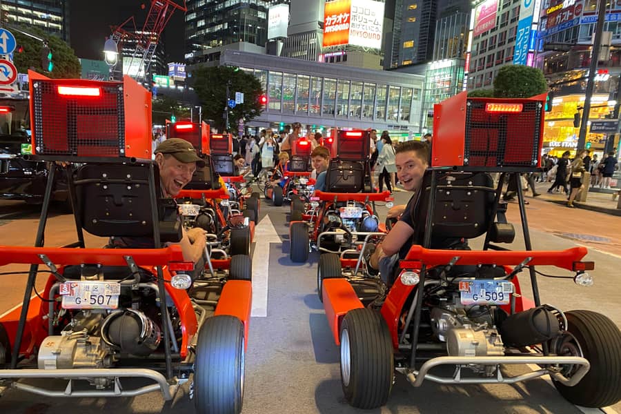 Night, red karts with rear cargo boxes lined up under neon signs, drivers seen from behind