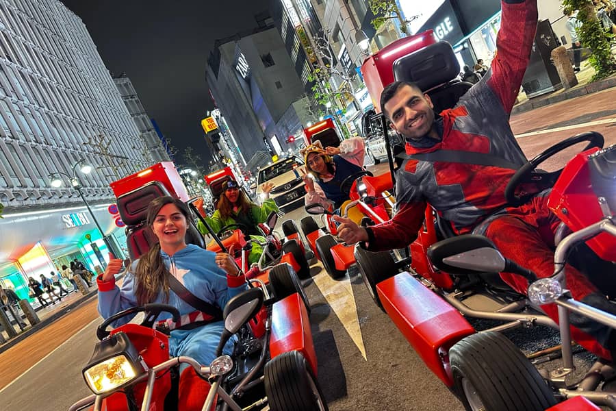 Night, woman in blue-green onesie and man in red at the wheel, neon Shibuya street, motion