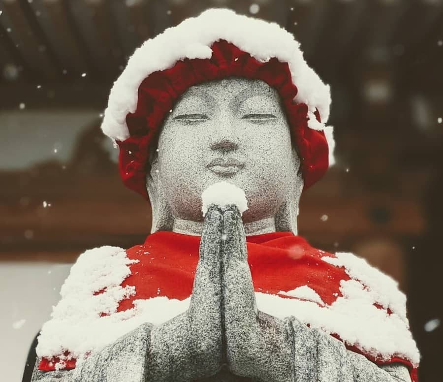 A stone Jizō statue with its hands pressed together, in a red knitted bonnet and bib, snow capping its head and falling around it
