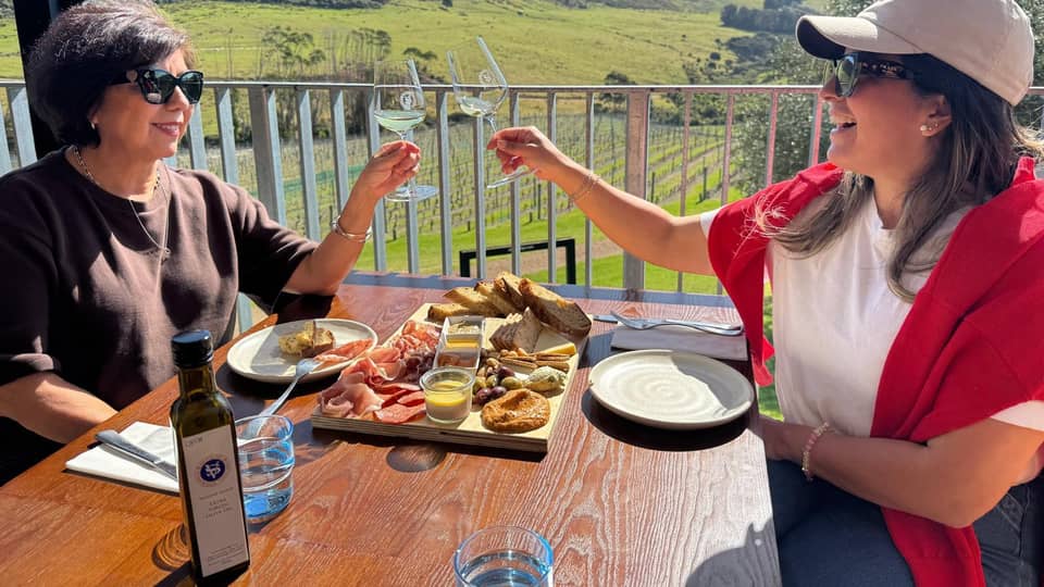 Two women sharing a platter and wine on a balcony over vines, close and warm