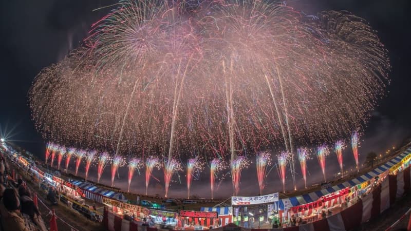 A wide fireworks burst filling the night sky above a curving row of lit festival stalls and the crowd below them