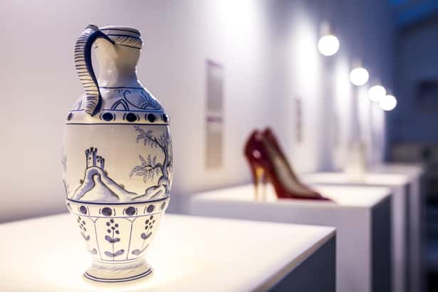 A decorative ceramic jug displayed on a pedestal in a museum, next to red high-heeled shoes, illuminated by soft lighting.