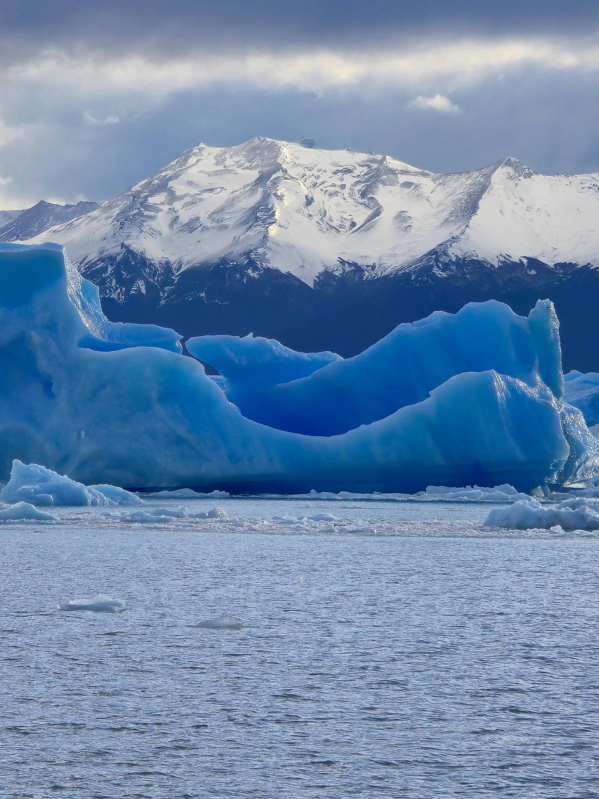 A deep blue iceberg in front of snowy peaks