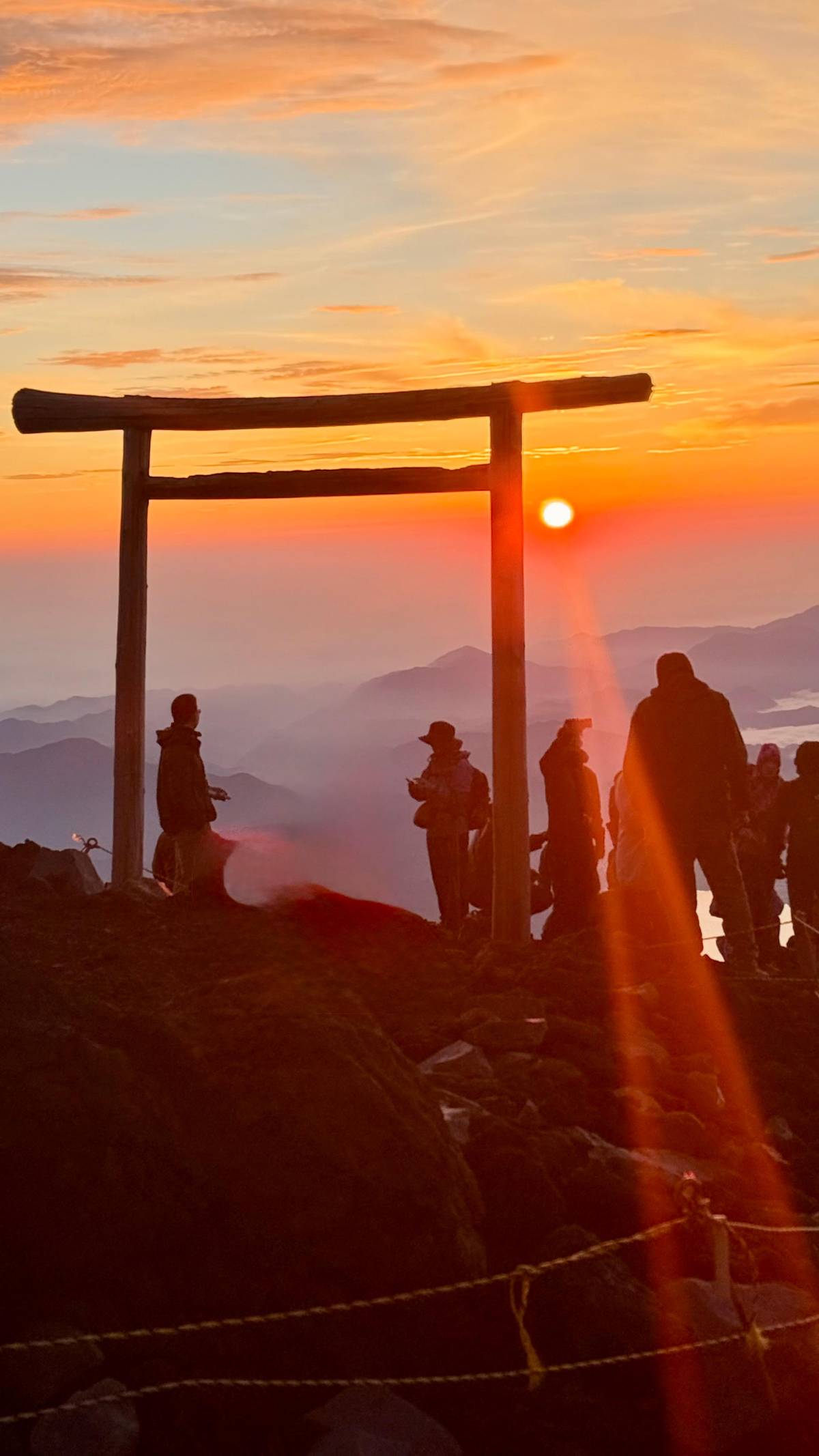 Climbers at a summit torii as the sun rises over the clouds