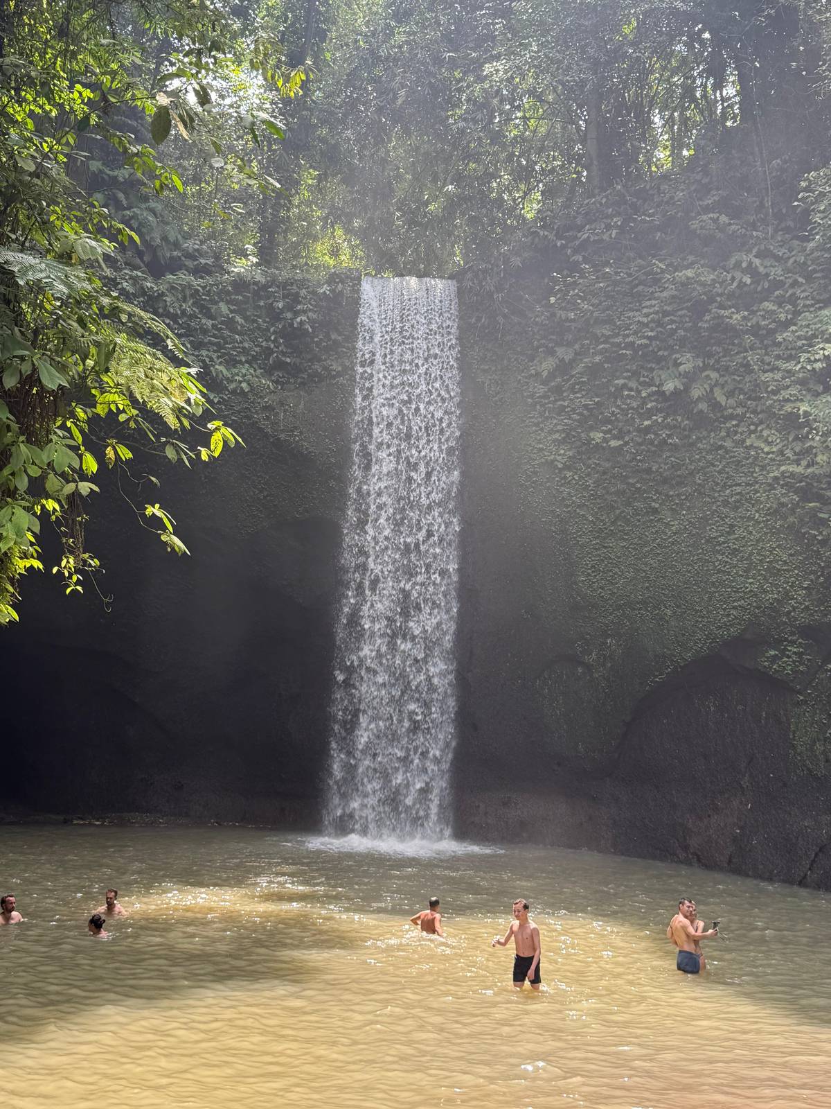 A tall waterfall dropping into a brown pool with swimmers