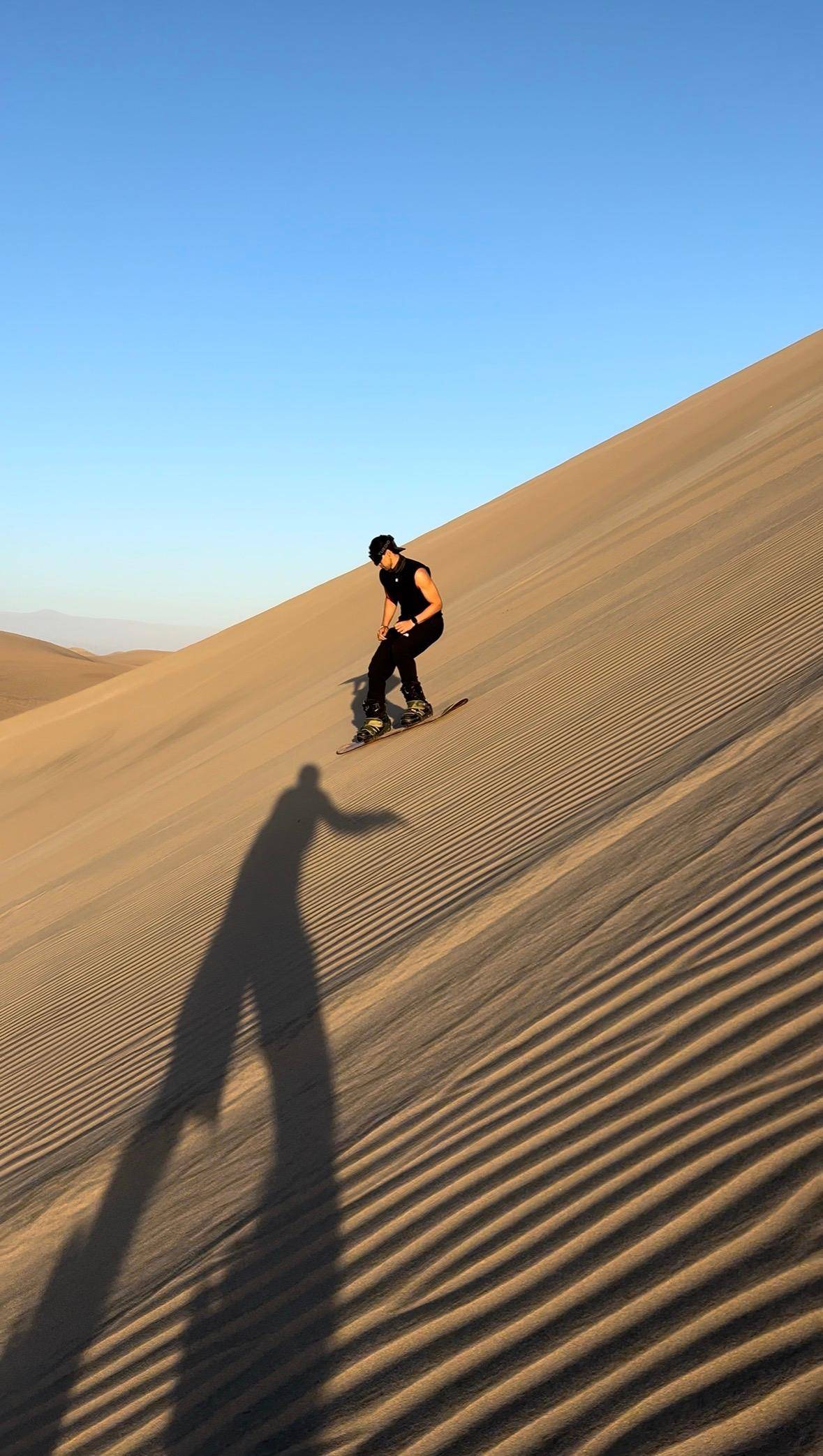 A sandboarder carving down a steep dune, his long shadow on the sand