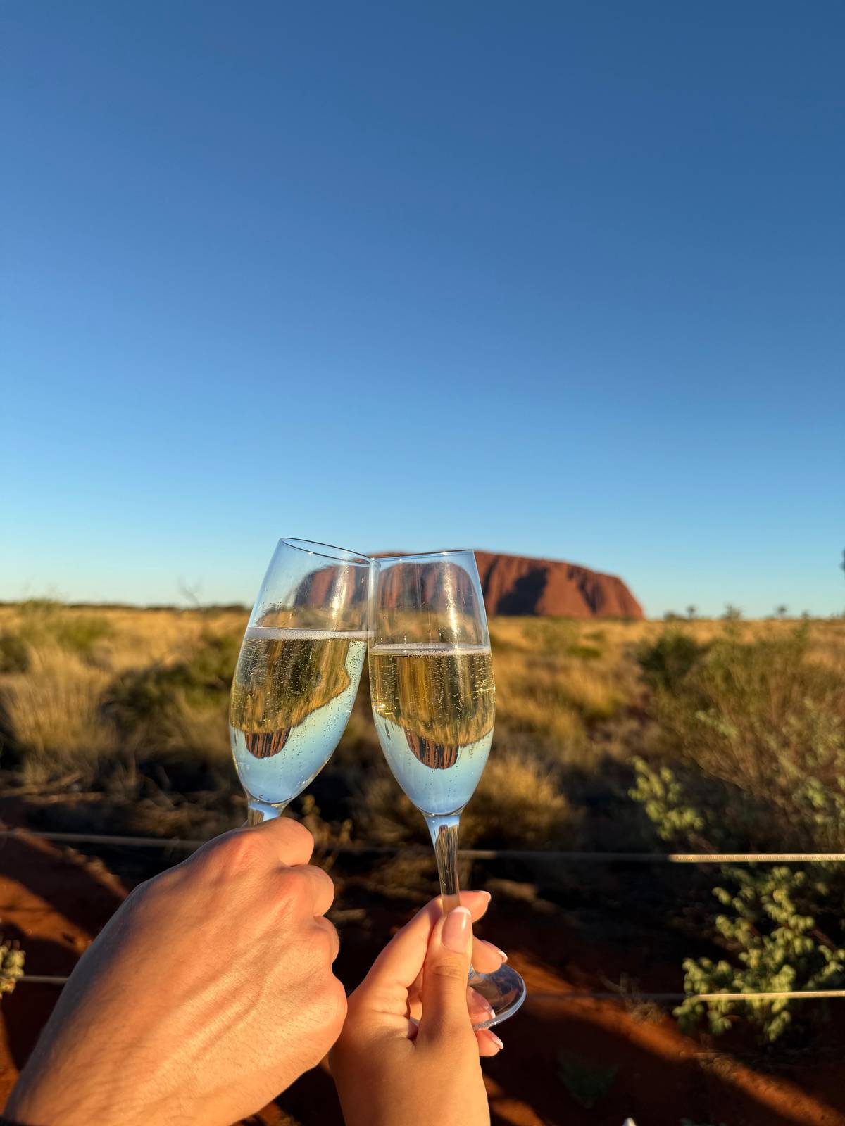Two glasses of sparkling wine raised with Uluru on the horizon