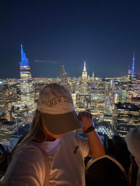 A visitor in a New York cap at the rail after dark, the lit skyline running south behind them
