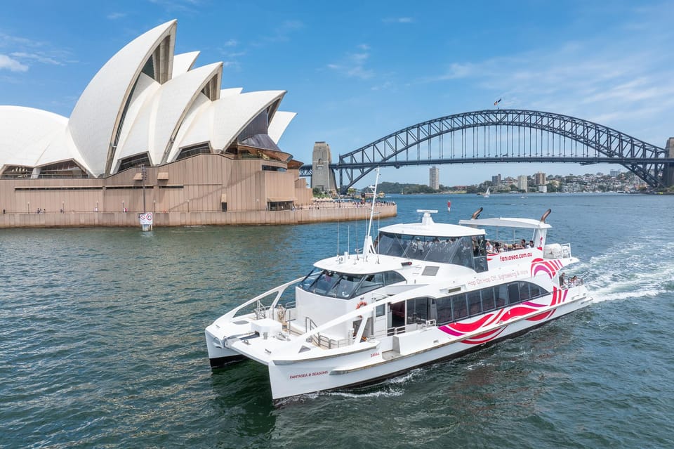 A white catamaran with a red swirl on the hull crossing open water, the Opera House sails on the left and the steel arch of the Harbour Bridge behind