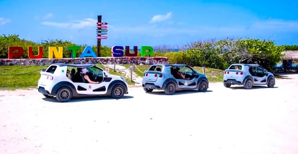 Three white buggies parked on pale sand in front of the colourful PUNTA SUR lettering, low scrub and blue sky behind