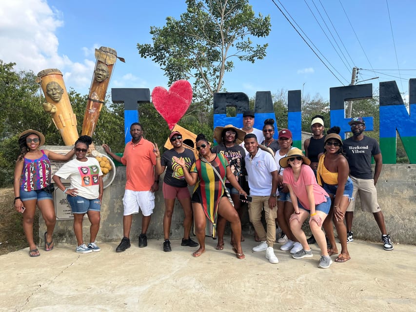 A tour group of about a dozen people posing under large painted letters and a red heart at a village entrance, two carved wooden posts with faces beside them, dry ground and a blue sky