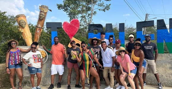 A tour group of about a dozen people posing under large painted letters and a red heart at a village entrance, two carved wooden posts with faces beside them, dry ground and a blue sky