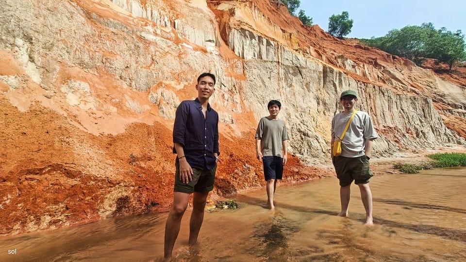 Three people standing ankle-deep in a shallow stream between orange and white layered sandstone walls