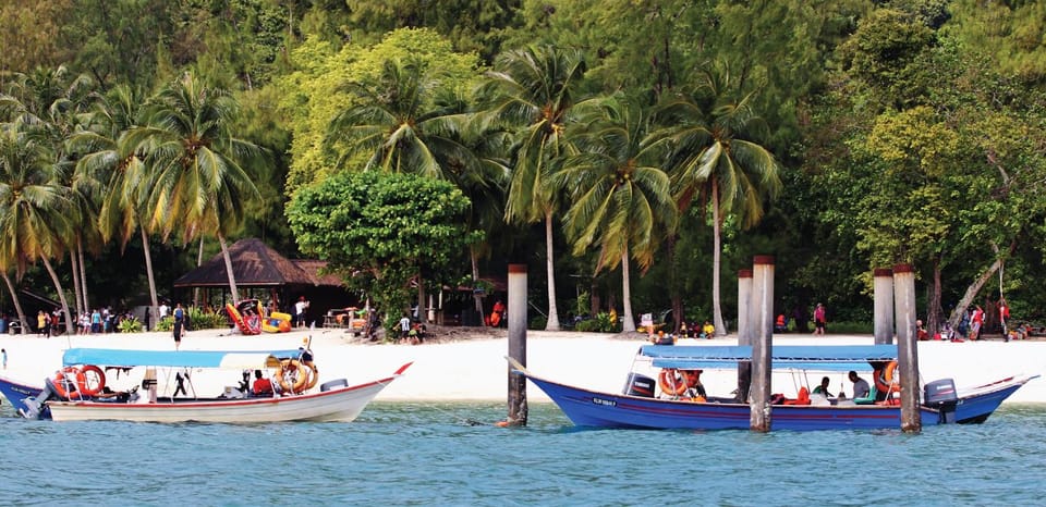 Two moored longboats in front of a white sand beach lined with coconut palms, people sitting under the trees on a bright day