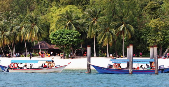 Two moored longboats in front of a white sand beach lined with coconut palms, people sitting under the trees on a bright day