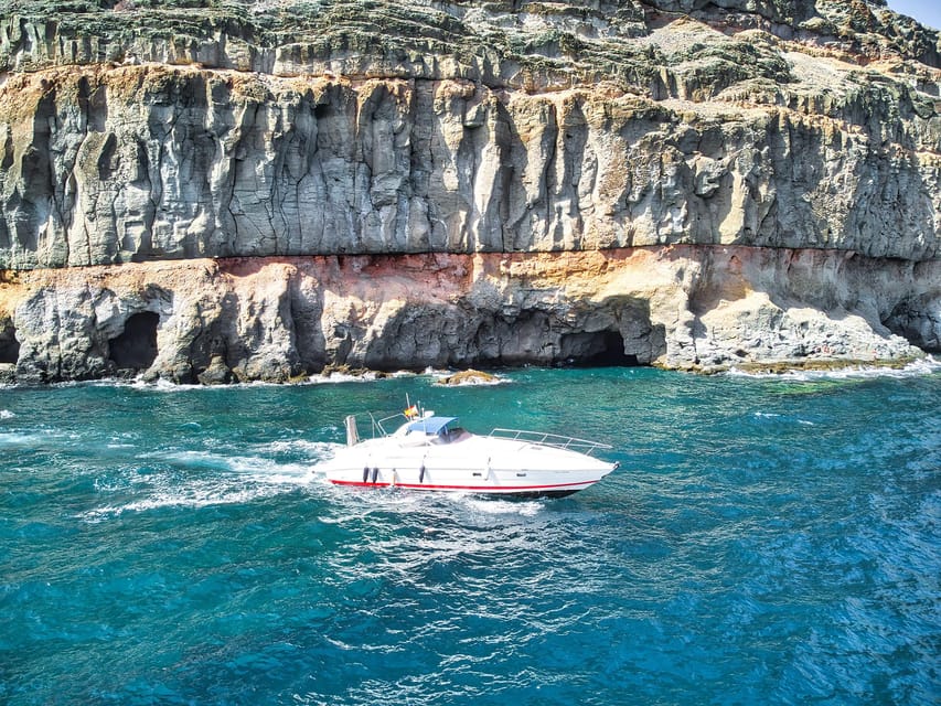A small white motorboat crossing dark blue water below steep brown volcanic cliffs
