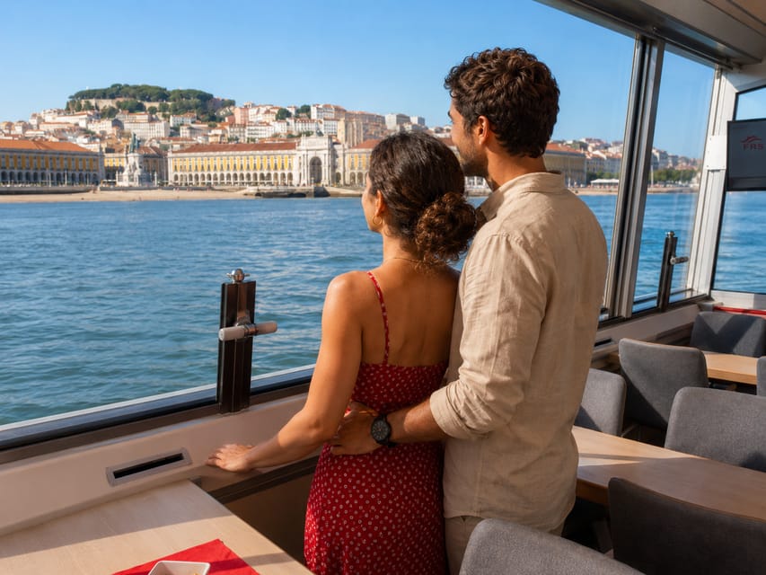 A couple standing at the window of a boat's glassed-in saloon, looking out at the Praça do Comércio and the castle hill behind it