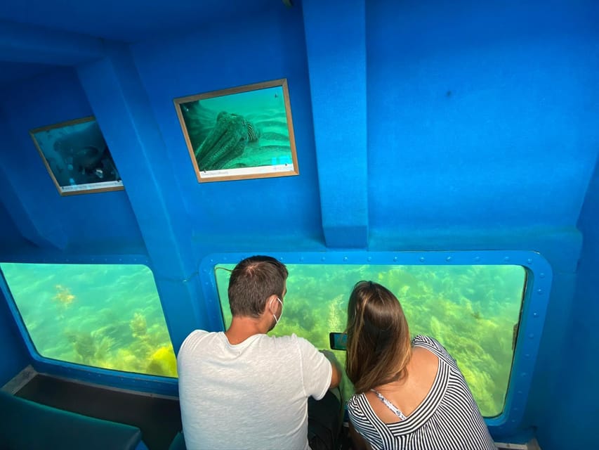 Two people looking through blue-framed underwater windows in a boat hull at green seabed