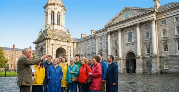 Guide gesturing to a group in raincoats on wet cobblestones in front of the Campanile and Georgian facade of Trinity College