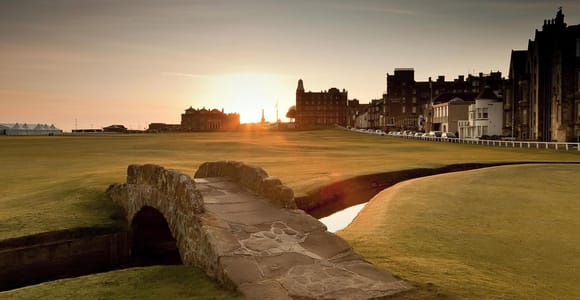 A small stone bridge over a narrow burn on a golf course at sunset, with the buildings of a town behind it