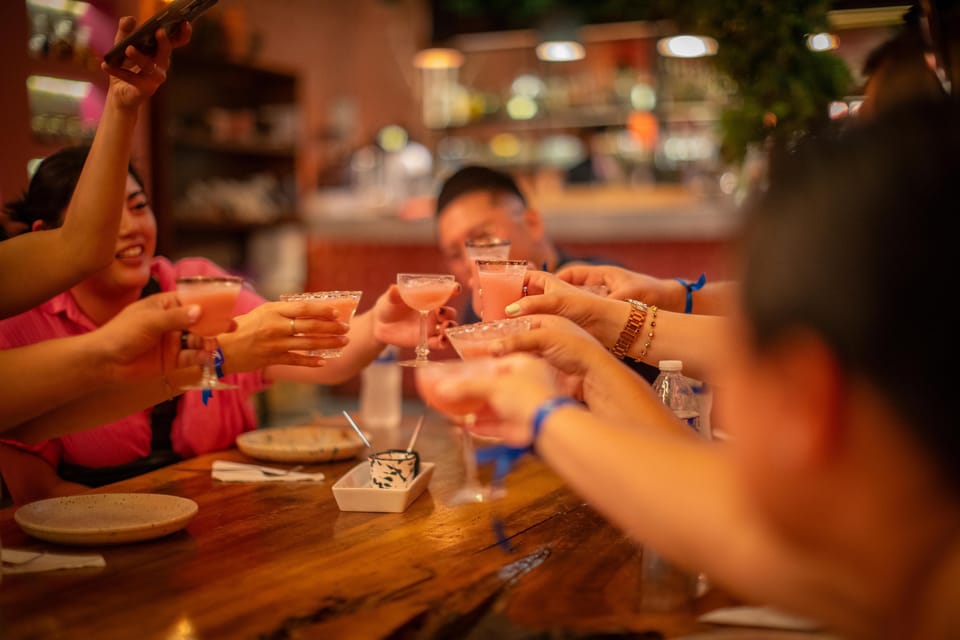Several hands reaching across a wooden bar to clink salt-rimmed cocktail glasses, warm lamplight and bottles blurred behind