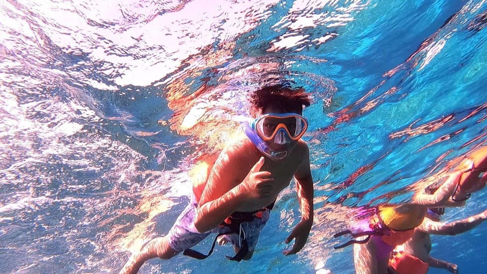 Underwater shot of a boy in an orange-framed mask and snorkel giving a thumbs-up, with other snorkellers to the right
