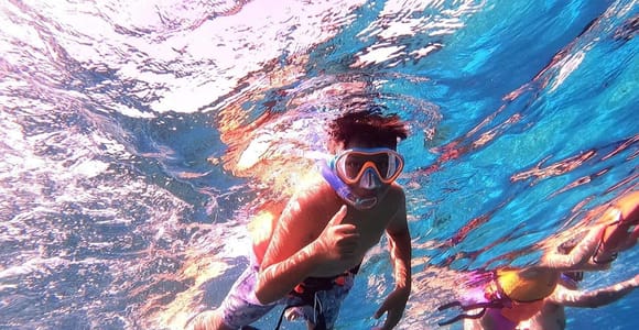 Underwater shot of a boy in an orange-framed mask and snorkel giving a thumbs-up, with other snorkellers to the right