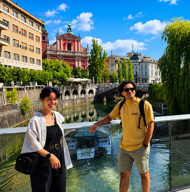Two travellers lean on a glass-fronted bridge in Ljubljana, with a pink-red church and a stone bridge over the river behind them
