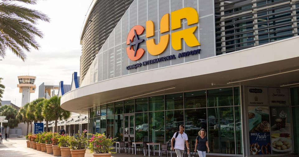 The kerbside frontage of Curaçao's airport terminal with large orange and yellow CUR letters above the entrance, palms in pots and two arriving passengers wheeling suitcases past the glass front