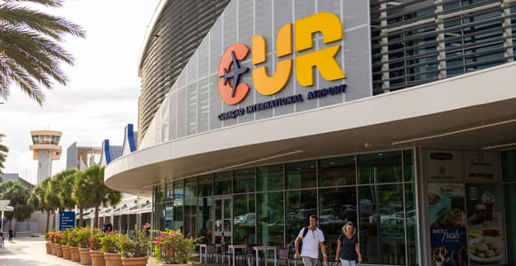 The kerbside frontage of Curaçao's airport terminal with large orange and yellow CUR letters above the entrance, palms in pots and two arriving passengers wheeling suitcases past the glass front