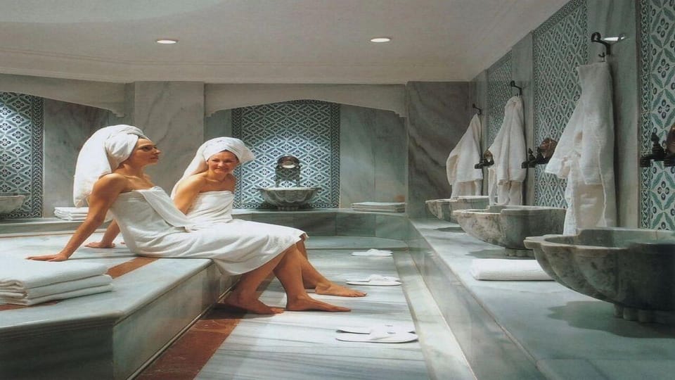 Two women in white towels and head wraps sitting on a marble bench in a tiled hammam with stone basins