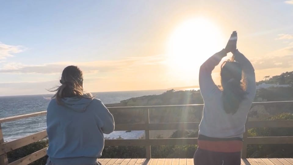 Two people practise yoga on a wooden clifftop terrace, facing the sea and the low sun