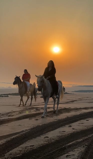 Two riders on horseback walk along a wide, wet beach at sunset, the low sun a bright disc in an orange sky behind them, tyre tracks in the sand