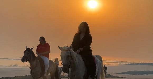 Two riders on horseback walk along a wide, wet beach at sunset, the low sun a bright disc in an orange sky behind them, tyre tracks in the sand