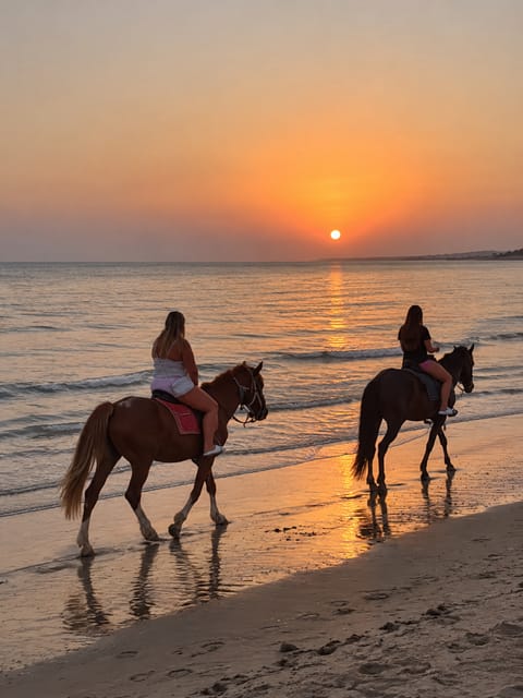 Two riders on horses at the edge of the sea on wet sand, with a low orange sun and its reflection on the water